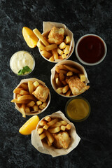 Paper cups with fried fish and chips, and sauces on black smokey background