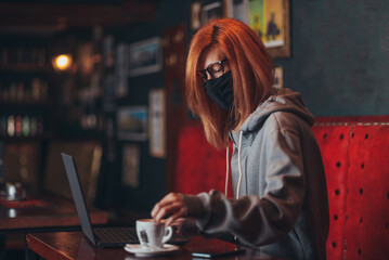 Woman with mask working on a laptop in a bar