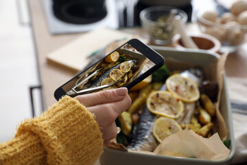 Female food photographer with mobile phone taking picture of tasty baked fish with potato in kitchen