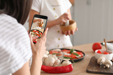Female food photographer with mobile phone taking picture of pizza maker in kitchen - obrazy, fototapety, plakaty