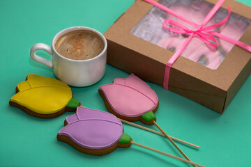 Gingerbread in the form of yellow. pink and lilac tulips with a white coffee cup and a festive box on a turquoise background. Festive food background.The concept of a holiday coffee break.