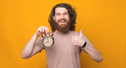 Portrait of joyful young bearded man with long hair showing thumb up gesture and alarm clock