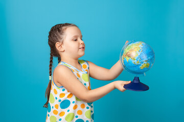 portrait of a cheerful girl in a white summer dress twirling a globe in her hand and choosing a country to travel, isolated on a blue background.