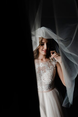 portrait of the bride. woman in a wedding dress stands near the window. the bride holds the veil. dark background. portrait of a beautiful girl.