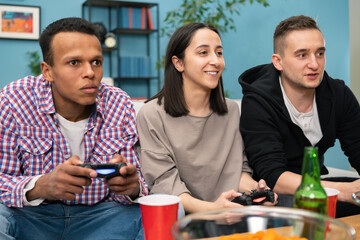 Multinational friends sitting at home on couch and playing video games on console. Cheerful woman having fun winning over man in game.