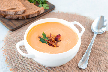 Pumpkin soup with fried bacon in a white plate with spoon served with bread and herbs near napkin on white table. Top view.