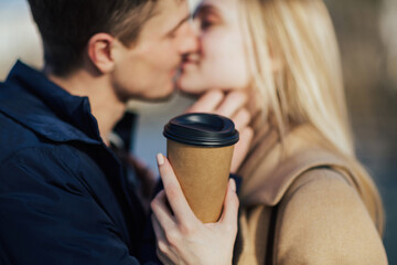 Close up of man and woman is kissing with  paper cup with hot drink in hand while walking in park.