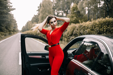 A beautiful young girl in a red overalls stands by a black car on an empty road in the forest