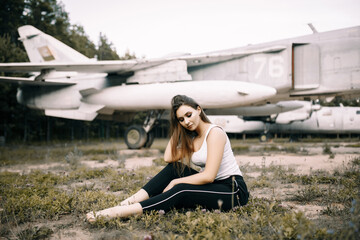 beautiful young brunette girl sits on the background of old military aircraft. Girl in a white shirt and black pants in nature. Military equipment. Portrait half-length. girl posing