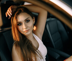Fototapeta na wymiar A beautiful young girl sits in a car in the evening in the sunset summer sun in an empty parking lot