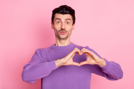 Photo of cheerful flirty young man dressed purple pullover showing heart sending kiss isolated pink color background