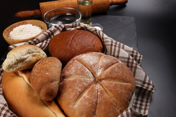 Freshly baked bread loaf bun roll round long mix verity wrapped in checkered kitchen fabric napkin towel wheat flower oil water salt eggs rolling pin slate stone over black background