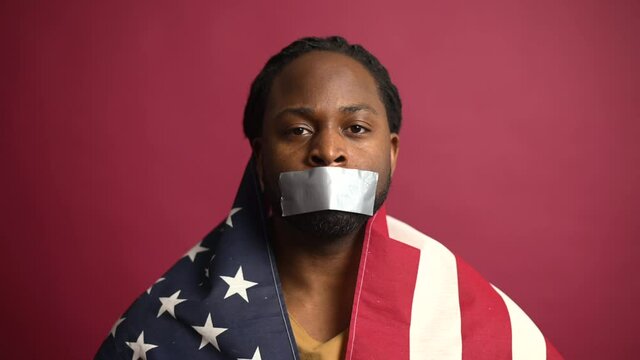 Confident African American fighter for rights voiceless speechless man covered with USA flag, mouth closed with adhesive tape, suffering from injustice, isolated on red wall, black lives matter