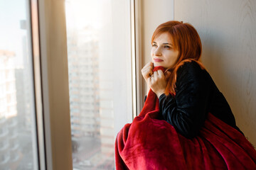 woman with red hair is sitting on a windowsill and wrapped with a duvet.