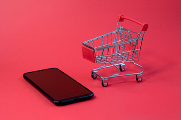 small shopping cart from supermarket on modern black smartphone close up on bright red background with blank space
