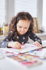 Cute child girl sitting at the table in earphones doing homework drawing using pencils or crayons. Homeschooling or art class concept. Vertical image. High quality photo