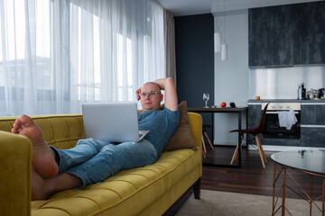 Man in a blue t-shirt lying on the yellow sofa and using laptop. Remote work, work from home.