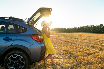 Young woman driver in yellow summer dress and straw hat standing near her car enjoying warm summer...