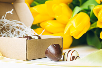 Chocolate candies in craft box and bouquet of yellow tulips on white wooden surface