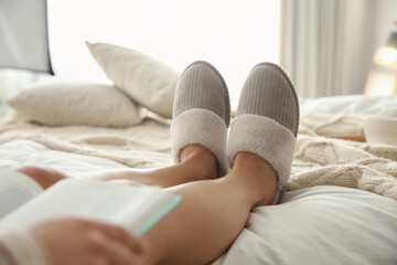Fototapeta na wymiar Woman in soft comfortable slippers reading book on bed at home, closeup