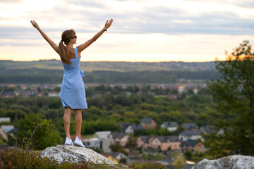 A young woman in summer dress standing outdoors with outstretched arms enjoying view of bright yellow sunset.