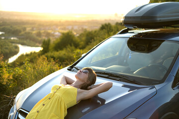 Happy young woman driver in yellow dress leaning on her car enjoying warm summer day. Travelling and vacation concept.