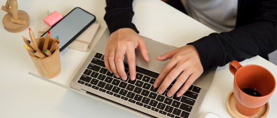 Female hands typing on laptop keyboard on home office desk with stationery, mug and smartphone