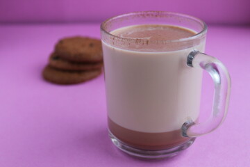 a cup of hot chocolate cocoa in a glass cup in the background lies homemade cookies and cinnamon sticks on a lilac purple background with a place for text and copyspace