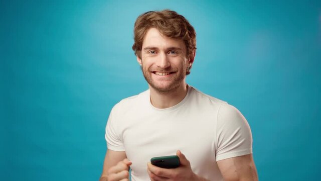 Young man looks at cell phone screen with joy, recieved good news, against blue background