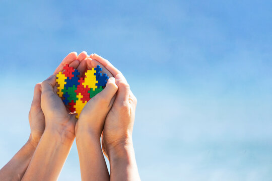 Autistic kid hands with multicolored puzzle heart supported by mother hands over blue sky background. World autism awareness day