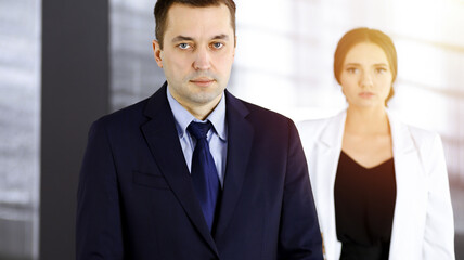 Portrait of a middle aged businessman in a dark blue suit, standing in a sunny office with a colleague. Business people concept