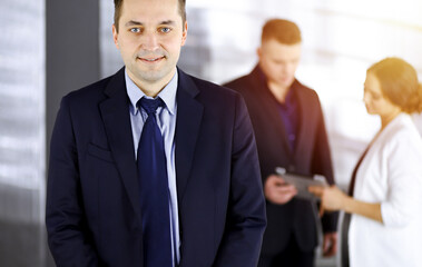 Portrait of a self-confident middle aged businessman in a blue suit, standing in a sunny modern office with his colleagues at the background. Concept of business success