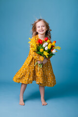 Cute little blonde girl holding a bouquet of tulips in a cotton dress on a blue background. spring, space for text