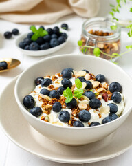 Yogurt with muesli, nuts and blueberries in a ceramic plate on the white kitchen table. Soft cream cheese with muesli and berries in a bowl closeup. Healthy Breakfast Concept