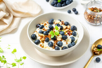 Yogurt with muesli, nuts and blueberries in a ceramic plate on the white kitchen table. Soft cream cheese with muesli and berries in a bowl closeup. Healthy Breakfast Concept