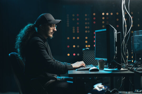 Middle aged Hackers with curly long hair in a sweatshirt, baseball cap and glasses making cryptocurrency fraud using virus software and computer interface. Blockchain cyberattack, ddos and malware