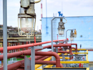 Row of lighting masts with retro design lanterns in explosion proof and fireproof design close-up over background of pipelines buildings and equipment of chemical plant with copyspace.