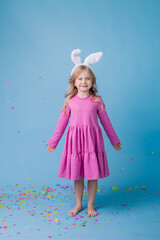 Fototapeta premium Cute little blonde girl in Easter bunny ears in pink dress on a blue background. Easter concept, text space