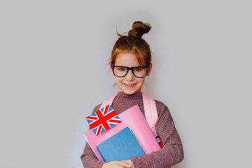 Happy girl kid 6 y.o. with black glasses holds a miniature of the English flag ,textbooks, and a school backpack. Learn English. Concept of education.