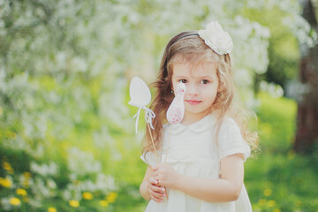 Happy Easter. Little girl in white dress hold easter toy eggs on sticks in the spring cherry garden. Portrait of happy child among white flowers trees. Childhood. Walking in sunny blooming park.