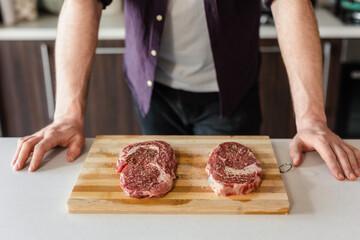Close Up Raw Steak Meat seasoned with Condiments, Salt and Pepper on a Wooden Board in Kitchen ready to be grilled by chef, man standing in background, copy space, food commercial photography