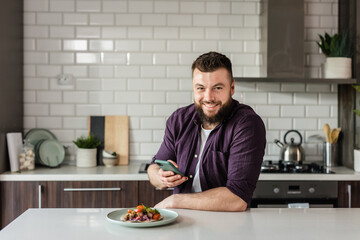 Fototapeta na wymiar Happy Man using Smartphone App Food Delivery and eating Dinner at Home in Modern Kitchen, Portrait, Copy Space