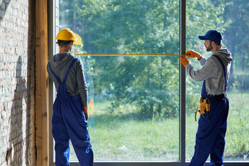 Fototapeta na wymiar Making new home. Rear view of two young male builders wearing blue overalls using measuring tape while working on cottage construction site