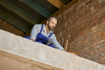 Young male builder in blue overalls looking enthusiastic while working on cottage house construction