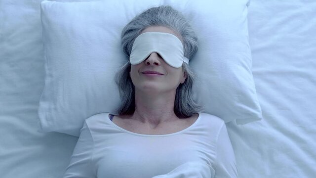 Smiling attractive woman lying in bed, wearing eye mask, healthy sleep habit