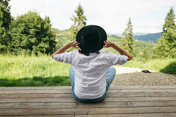 Young female in hat and casual cloth sitting on porch on background of sunny mountains hills. Travel