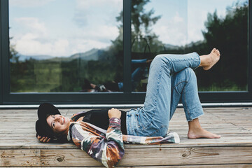 Stylish happy woman relaxing on wooden terrace at modern cabin with windows in mountains. Travel