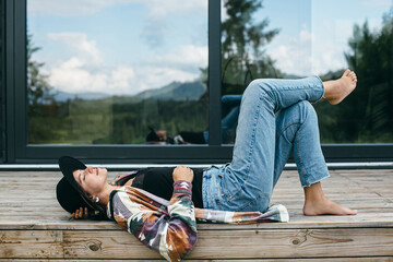 Stylish happy woman relaxing on wooden terrace on background of modern cabin in mountains. Tranquil