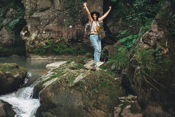 Happy woman traveler with backpack standing on rocks of river in mountains,  holding hands up