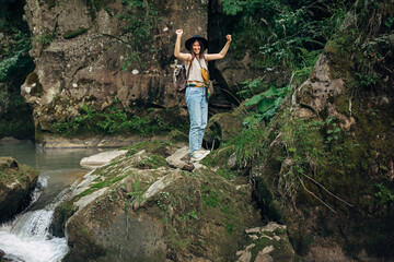 Obraz premium Happy woman traveler with backpack standing on rocks of river in mountains, holding hands up